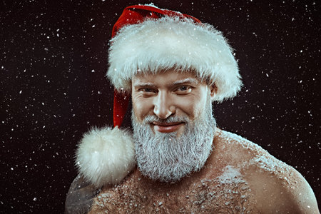 A brutal Santa Claus with an athletic build stands in a cap with bared torso and smiles. Black background with snow and space for text.の写真素材