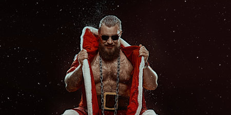 Christmas and New Year concept. A handsome brutal muscular Santa Claus in a red fur coat and sunglasses stands covered in snow and looks at the camera. Black background with snowfall. Place for text.の写真素材