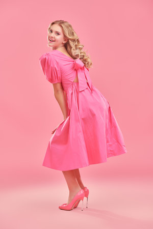 A beautiful slender girl with long curly blond hair poses flirtatiously in an elegant pink dress and high-heeled shoes. Full length portrait on a pink studio background. Women's beauty and fashion.の写真素材