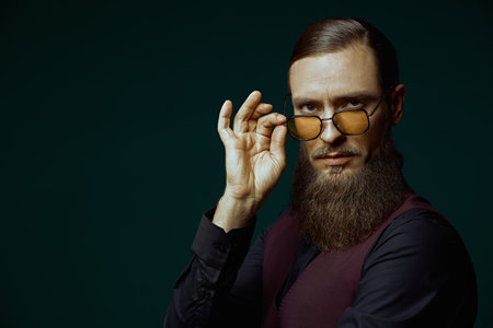 A respectable, well-groomed mature man in an elegant black shirt and vest looks seriously at the camera through his glasses. Black background with copy space. Businessman.の写真素材