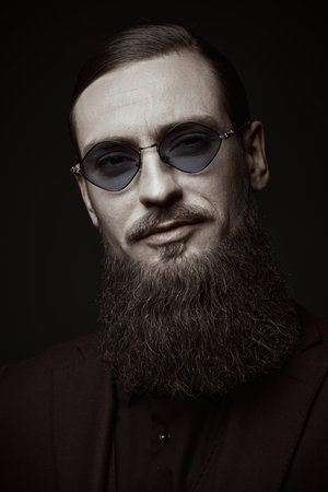 Portrait of an imposing mature man with a long beard, dressed in an elegant suit and dark glasses, smiling at the camera. Black studio background. Men's beauty, beard and hair style.の写真素材