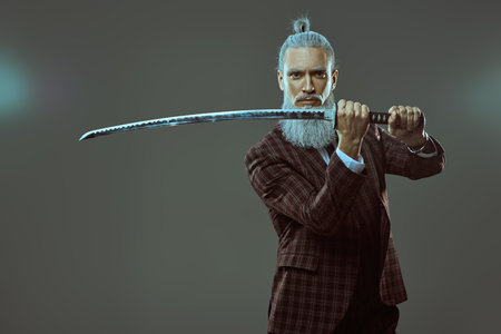 A gray-haired mature man in an elegant suit poses with a samurai sword. Japanese style. Business and determination. Gray studio background with copy space.の写真素材