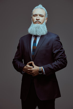 Style and fashion for men. A handsome mature man with a gray hairstyle and a beard, dressed in an elegant suit, looks calmly and confidently into the camera. Gray studio background. Business concept.の写真素材