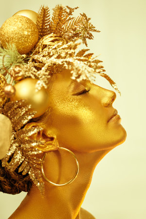Holiday luxury look. Christmas beauty portrait. Beautiful young woman with golden make-up and shiny golden skin poses in a Christmas wreath. White studio background. Close-up shot.の写真素材