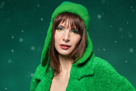 An attractive brunette young woman model posing in a bright green fur coat and green knitted hat on a green studio green background. Winter Christmas fashion style.の写真素材