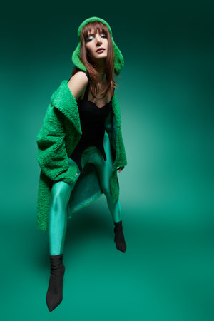Winter Christmas fashion style. Full-length portrait of an attractive brunette young woman model posing in a bright green fur coat, green knitted hat and leggings on a green studio background.の写真素材
