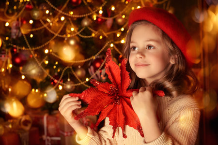Vintage fabulous Christmas and New Year. Portrait of a happy little girl in a red beret standing with a large red flower against the background of a Christmas tree with golden lights. Copy space.の写真素材