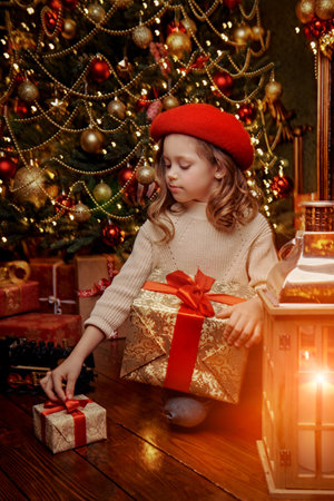 A cute little girl in a red beret sits with gifts on Christmas Eve next to a beautiful Christmas tree under the light of a lantern. New Year. Retro style.の写真素材