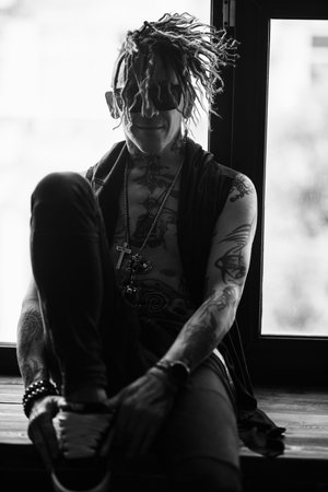 A brutal male rock musician dressed as a fallen black angel sits on the windowsill. Rock style.の写真素材