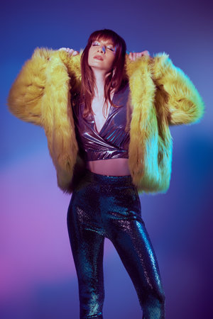 An attractive girl in a shiny top and leggings and yellow fur coat poses in a mixed neon light. Fashion and sparkle. Fashionable style for a glittering party.の写真素材