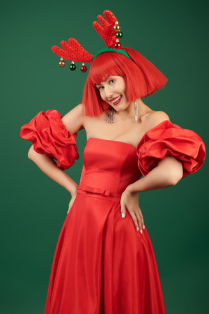 Celebration of Christmas and New Year. A pretty Asian girl model in an elegant red dress, a red wig and deer horns poses joyfully against a green background.の写真素材