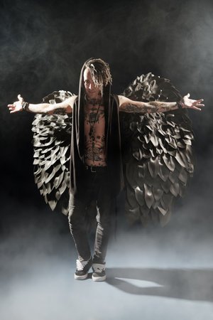 Full-length portrait of an artistic rock musician dressed as a fallen black angel spreaading his hands. The heavenly light.の写真素材