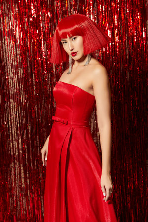 Evening festive fashion. Charming Asian girl model posing in an elegant long red dress and red wig against the background of shiny foil curtains. Christmas and New Year.の写真素材