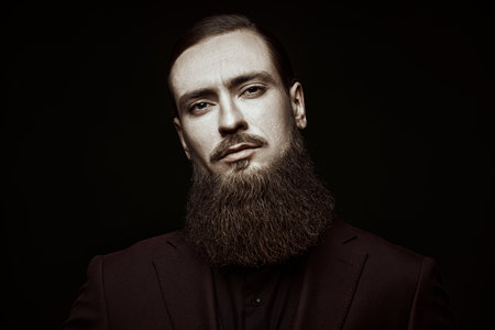 A portrait of a mature man with a long beard, dressed in a classic suit, looking thoughtfully to the camera. Black studio background with copy space. Business. People and characters.の写真素材