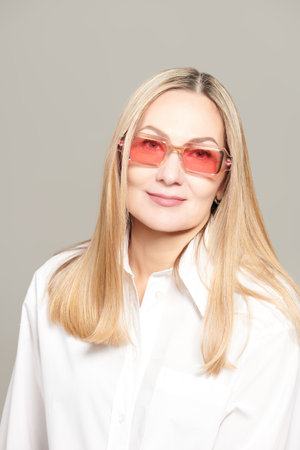 Portrait of a beautiful mature woman in a white blouse and fashionable glasses. Light gray studio background. Business style and fashion.の写真素材