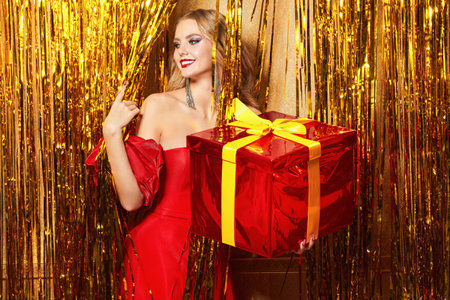 Evening makeup and hair styling. A pretty blonde lady posing in an elegant red dress with a golden gift box in her hands. Background with golden foil curtains.の写真素材