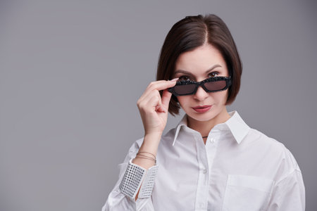 Glasses advertising. Portrait of a beautiful middle-aged woman in a white blouse and fashionable sunglasses. Light gray background with copy space. Business style and fashion.の写真素材