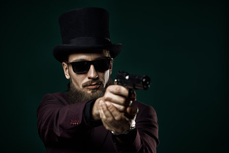 Crime, mafia. An elegant man with a long beard, dressed in an three-piece suit and a top hat, poses with a gun against a black background. Retro style. Men's fashion. Copy space.の写真素材