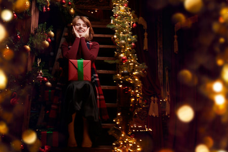 A joyful red-haired girl sits on wooden steps in a fairytale Christmas setting with a gift on her lap. Golden lights all around. Place for text.の写真素材