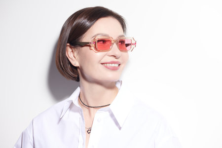 Glasses fashion. Happy mature woman in a white blouse and transparent pink sunglasses smiles, bathed in light on a white background. Light summer style. Copy space.の写真素材