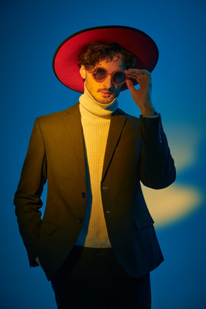 Men's fashion, clothing and accessories. Portrait of a stylish brunette man, fashion model, posing in a suit, wide-brimmed hat and round glasses. Studio shot in mixed color lighting.の写真素材