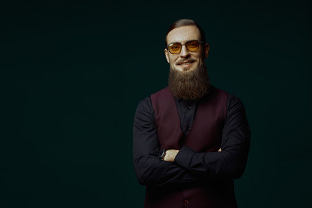 A respectable, well-groomed mature man in an elegant black shirt and vest looks seriously at the camera through his glasses. Black background with copy space. Businessman.の写真素材