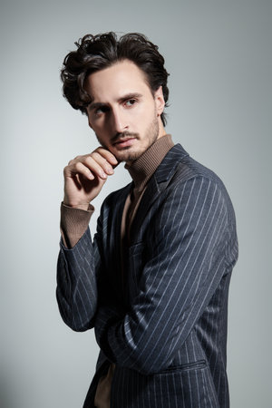 Men's beauty and style. Portrait of a handsome brunette man with curly hair in a turtleneck and a classic business suit. Men's hairstyles. Studio shot on a gray background.の写真素材