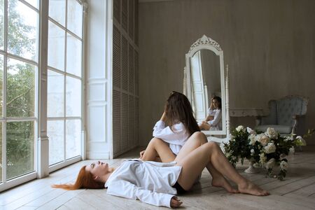 Two girls - a brunette and a redhead are sitting on the floor in a beautiful room. Amazingly beautiful girls in white shirts.の写真素材