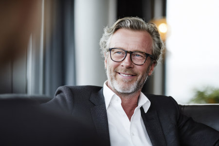 A mature man with graying hair and a beard, wearing glasses and a dark suit with an open white shirt, smiles as he looks to the side. He is seated indoors, with a blurred background that hints at a modern interior. His expression is relaxed and confident, conveying a sense of approachable professionalism.の素材