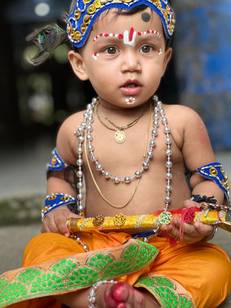 A close-up portrait of an adorable baby dressed as the Hindu deity Lord Krishna. The baby wears an orange dhoti, a blue headband adorned with a peacock feather, and various necklaces and armbands. Intricate forehead markings in white and red complete the divine costume. The baby holds a decorative flute.の写真素材