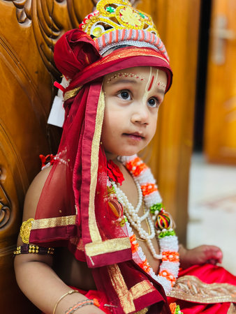 A close-up portrait of a young child dressed as the Hindu deity Krishna. The child wears a traditional Indian costume with a red dhoti, a maroon and gold turban topped with a crown, and a multi-colored garland. The child's gaze is directed to the side, conveying a thoughtful or pensive mood. The ornate wooden door serves as the backdrop, highlighting the rich details of the costume and the child's delicate features.の素材
