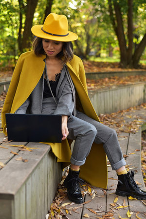 Asian business woman in a yellow coat sitting in the park and using a laptop. Autumn.