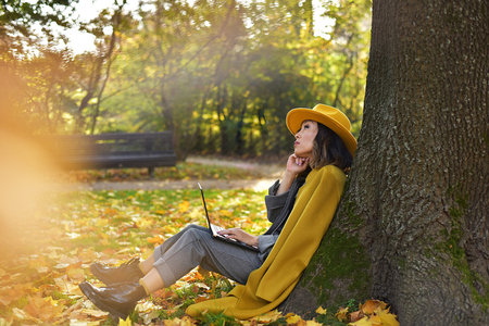 Asian business woman in a yellow coat sitting in the park and using a laptop. Autumn.の写真素材