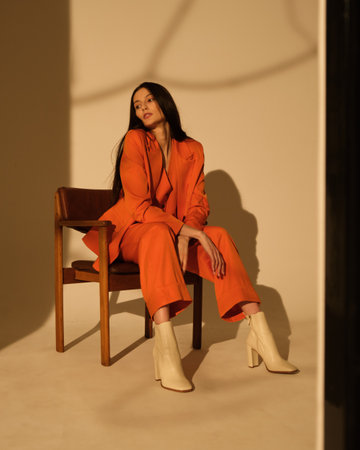 A beautiful young girl in an orange suit sits in a chair on a white background. Hard light.の写真素材