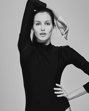 Black and white portrait. A beautiful young girl in a black long-sleeved bodysuit sits on a chair...