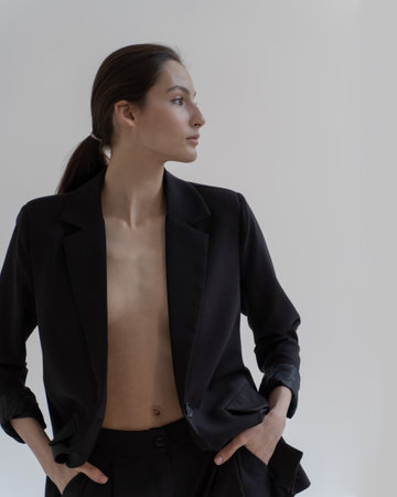 A beautiful young girl in black trousers and jacket on a naked body stands on a white background. Looks to the side.の写真素材