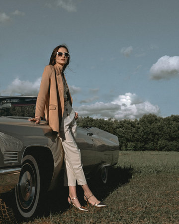 A beautiful woman stands next to a convertible in a field. Dressed in a beige jacket and white...