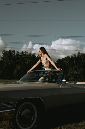 A beautiful woman stands in a convertible in a field. Dressed in white pants, a white top, and black...