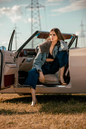 A beautiful woman sits on a convertible in a field. Dressed in jeans and a fur jacket. Beautiful and stylish look.の写真素材