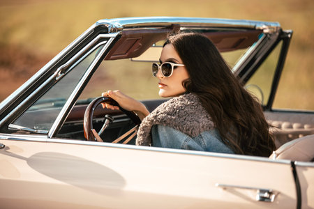 A beautiful woman sits on a convertible in a field wearing sunglasses. Dressed in jeans and a fur jacket. Beautiful and stylish look.の写真素材