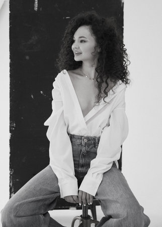 Black and white image. A beautiful young girl with curls in blue jeans and a white blouse sits on a bar stool on a black and white backgroundの写真素材