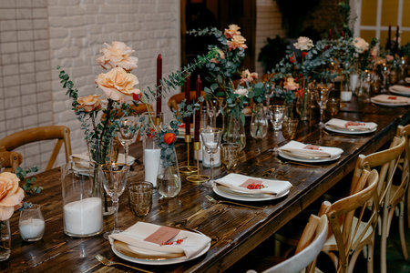 A beautifully set wedding table features elegant floral centerpieces with roses and eucalyptus, complemented by candles and stylish dinnerware on a rustic wooden surface.の写真素材