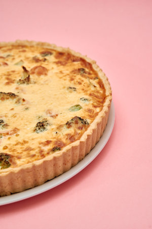 A savory broccoli cheese quiche rests on a white plate against a pastel pink background,...