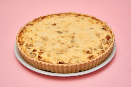 Golden-brown quiche lorraine sits on a white plate, featuring a flaky crust and creamy filling, set...