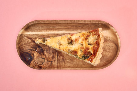 A slice of cheese and broccoli quiche rests on a wooden platter against a soft pink background,...