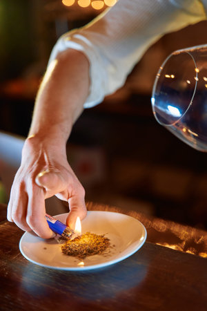 A bartender uses a lighter to ignite dried citrus peels, releasing aromatic smoke to enhance a...