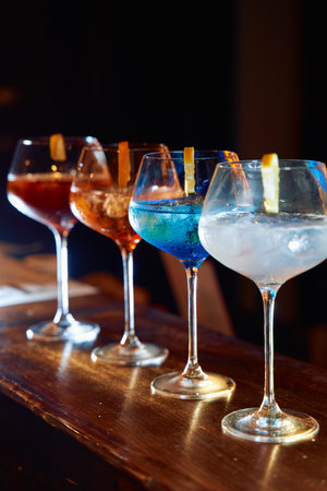Four colorful gin and tonics sit elegantly on a wooden bar, each with a distinct hue and citrus...