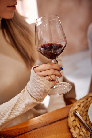 A woman in a beige sweater elegantly holds a glass of red wine at a rustic dinner setting, creating...