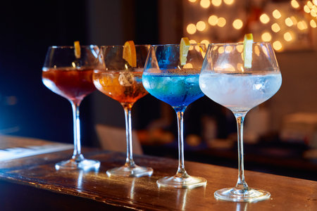 A lineup of four vibrant cocktails in elegant glassware, each garnished with lemon, sits on a bar...
