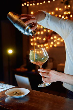 A bartender skillfully pours a cocktail from a shaker into a glass in a dimly lit bar with string...